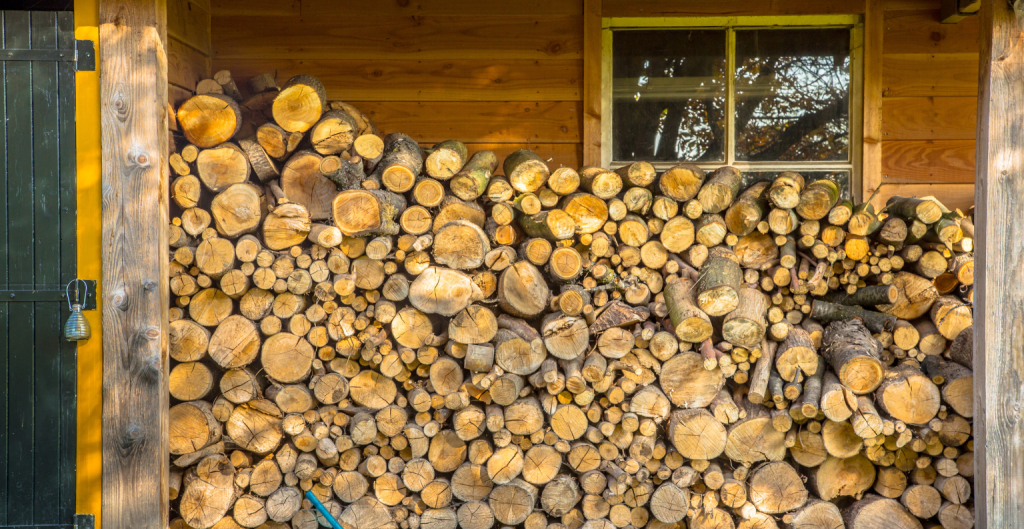 Split firewood logs stacked in a covered log store against a timber cabin wall, seasoning for winter